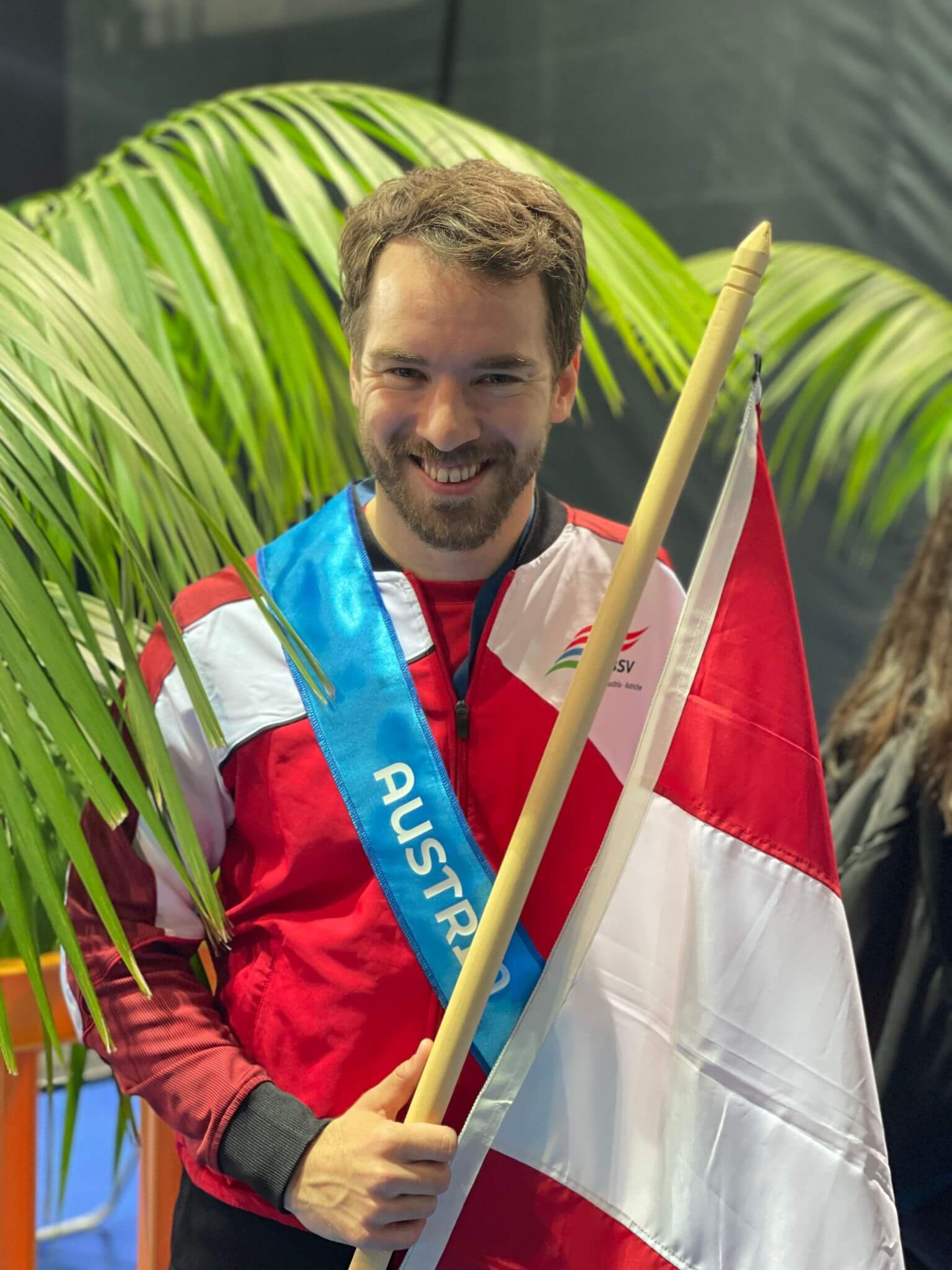 Andreas Onea im rot-weißen Trainingsanzug vor einer Zimmerpalme mit einer blauen Schleife um, auf der "Austria" steht, und der österreichischen Flagge in der Hand