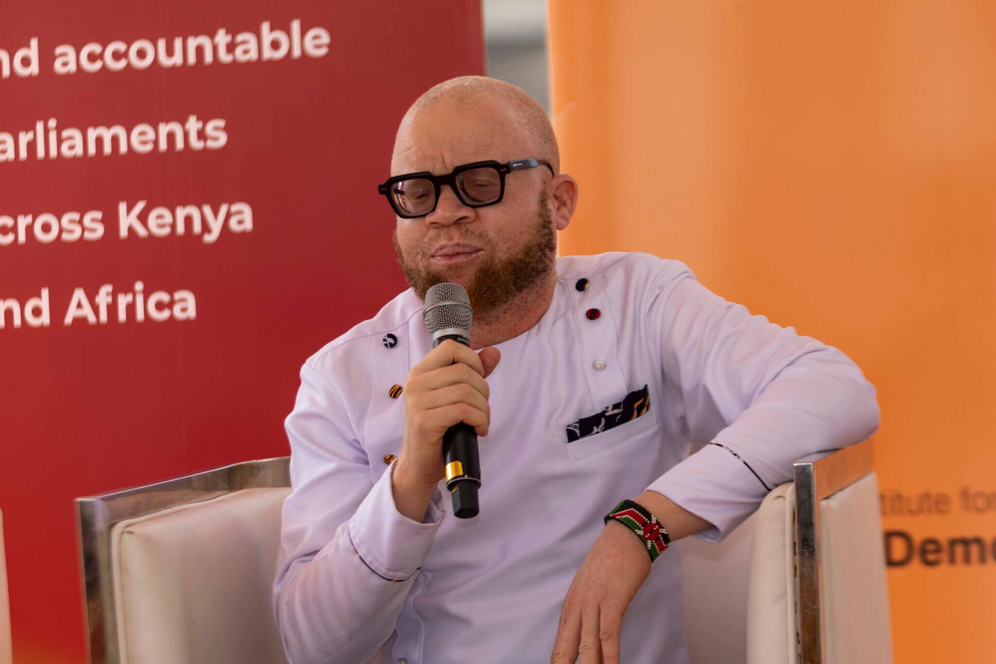 Collins Ombajo, a Disability Inclusion Advisor with Light for the World, dressed in white and holding a microphone. Collins is speaking at a panel discussion on "Walking the Talk on Inclusion of Persons with Disabilities in Electoral Processes".