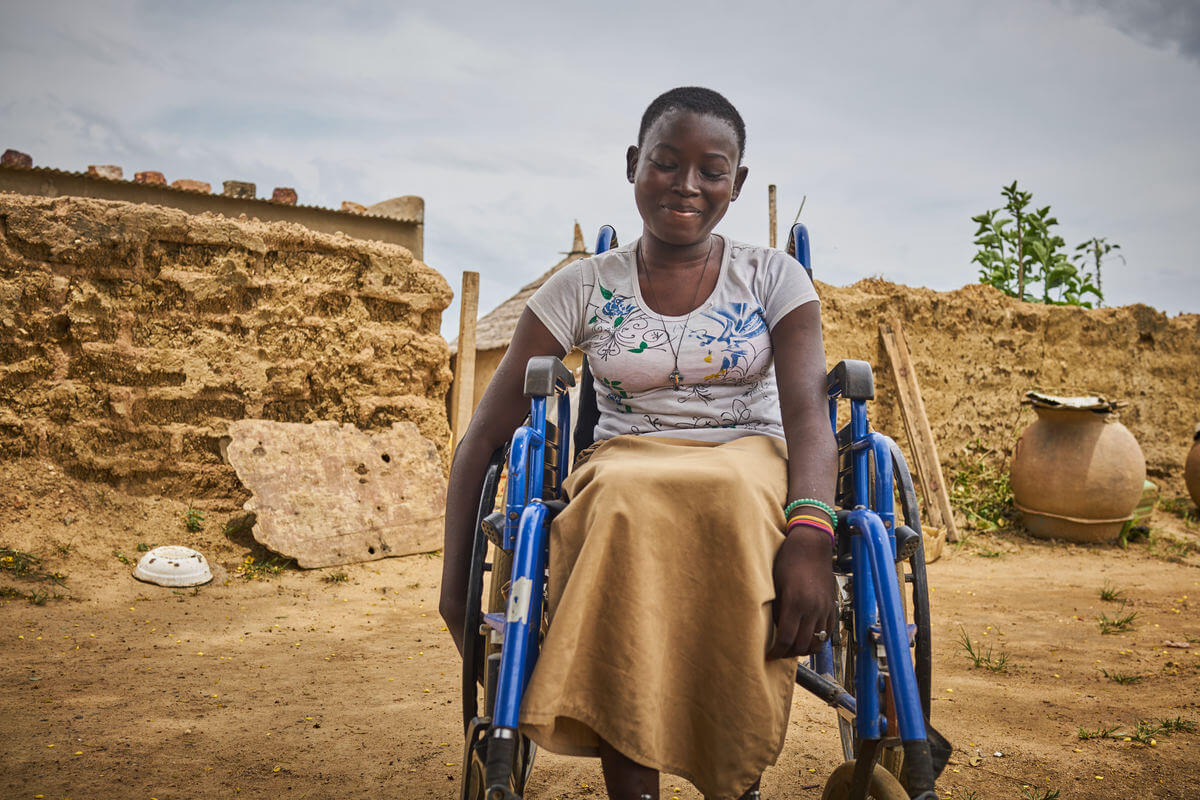 Philoméne, eine junge Frau, in ihrem blauen Rollstuhl, den Blick zum Boden gerichtet, lächelnd