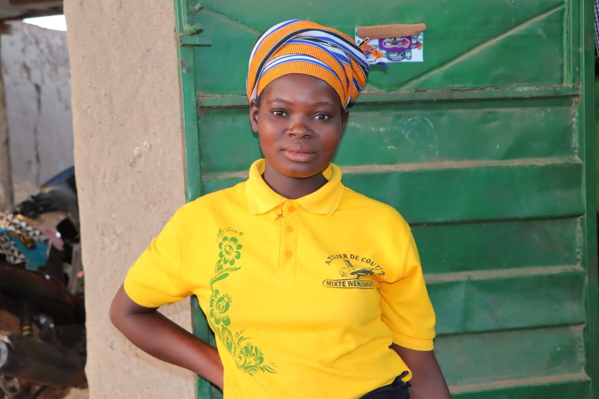 Karfo Gnonka in der Werkstatt, in der sie die Ausbildung zur Schneiderin macht. Sie trägt ein gelbes T-Shirt und hat die rechte Hand in die Hüfte gestützt.