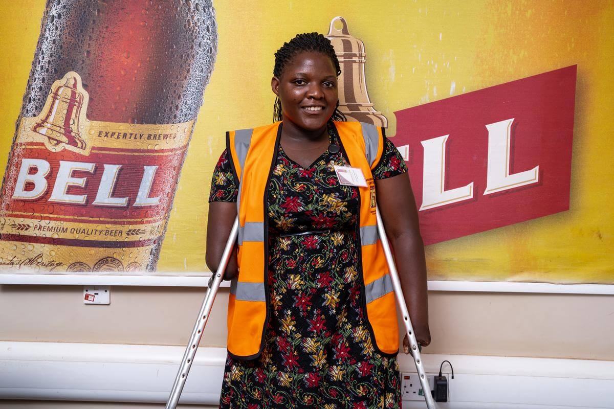 Christine Namata, Absolventin des Bachelor of Business Administration der Makerere University, steht vor einer Werbewand der Uganda Breweries Limited. Sie trägt eine orange Warnweste, ihre Arbeitskleidung.