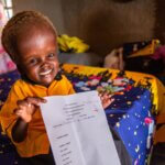 Nyamush is a little girl living with hydrocephalus and other physical disabilities. The girl is sitting on a bed in an IDP Camp and is showing her school test where she scored 100%. She has a big smile in her face and wears a yellow school uniform.