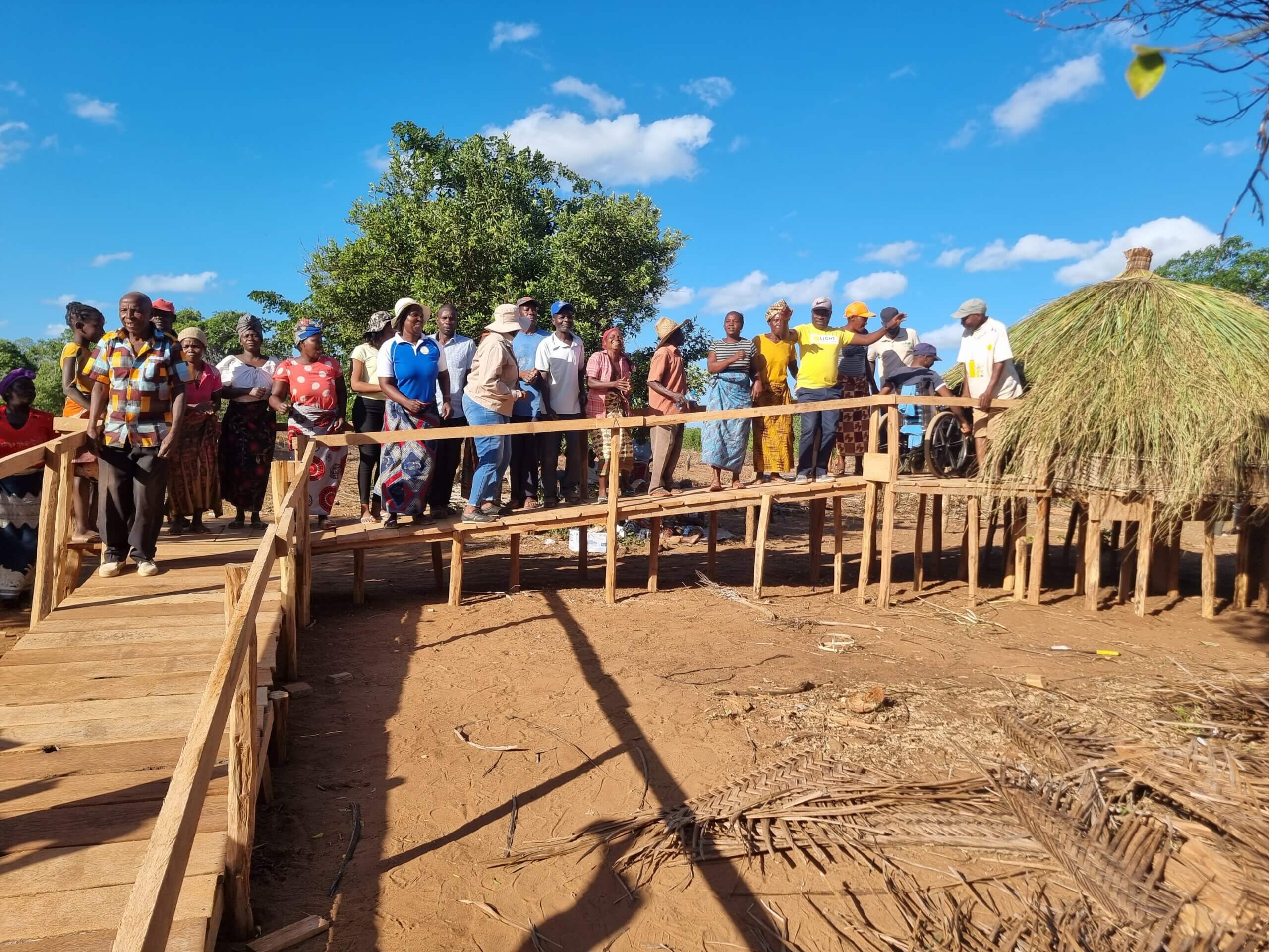 A group of people stand on a wooden ramp, leading to a newly constructed storage barn. It is covered in straw grass. The whole construction is on wooden stilts.