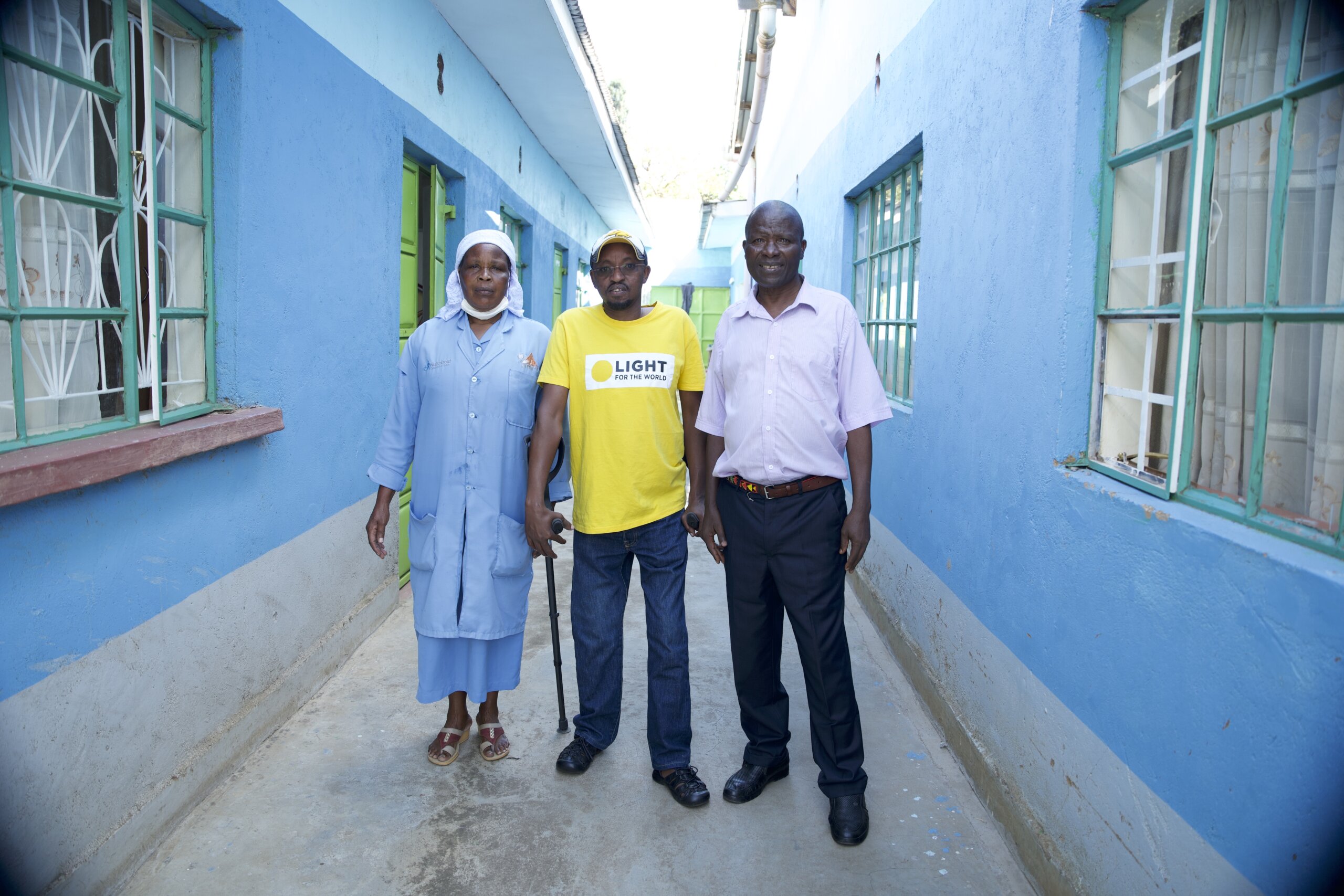 David Ndungu stands next to Peris and Stanley, both members of United Disabled People of Laikipia group. David is in the middle wearing a yellow Light for the World t-shirt and hat. A woman on his left wears a long light blue coat and skirt, and a white head scarf. The man on the right wears a pale shirt and dark trousers. The stand outside in front of bright blue walls.