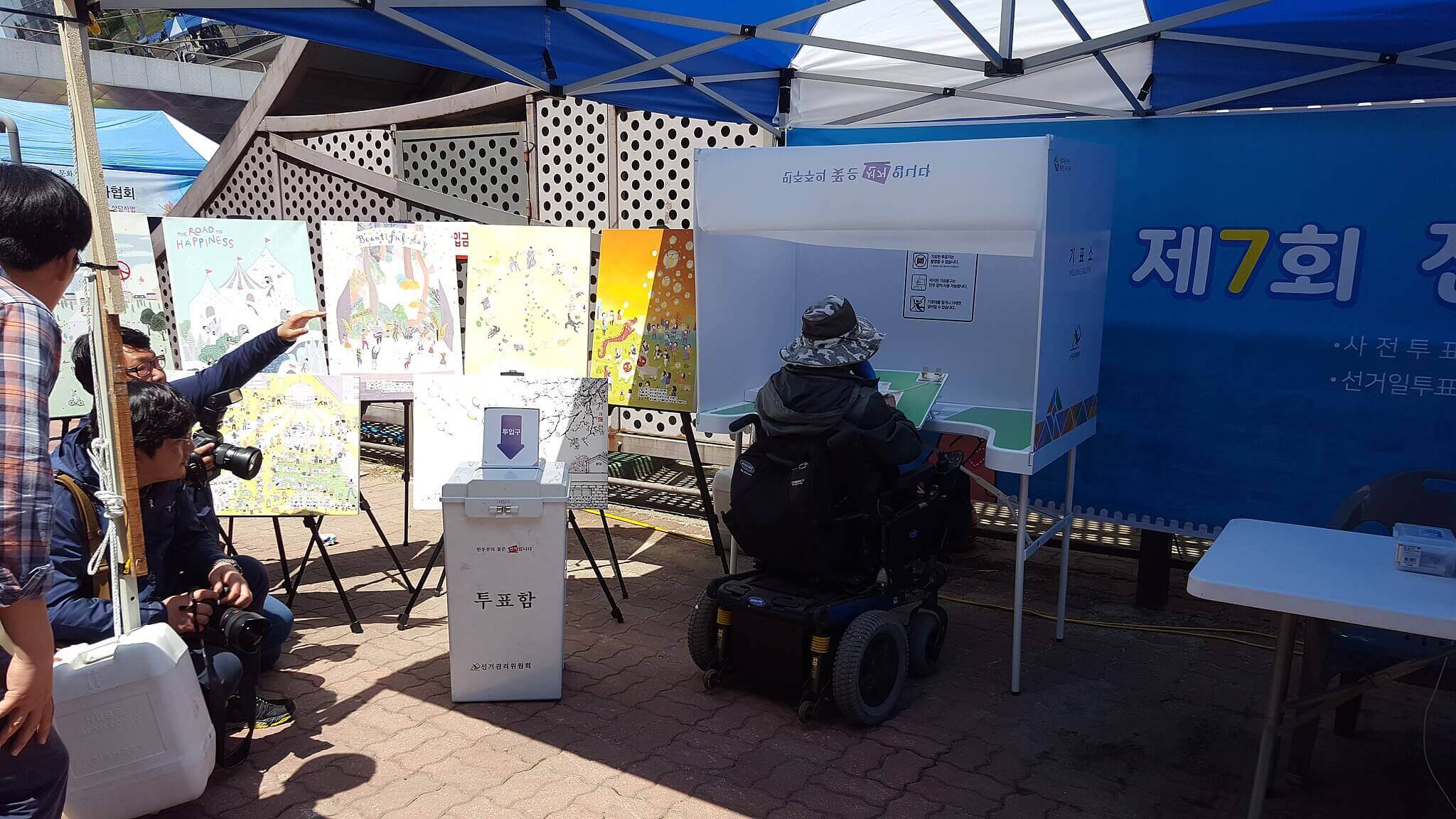 A wheelchair user votes at an accessible polling station in a mock election experience centre in South Korea. Journalists and a photographer are watching.