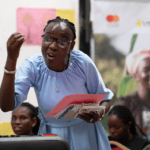 A female trainer stands and shares learnings with the programme participants. She wears glasses, a light blue dress, and holds note books in her hands. Her expression is engaging.