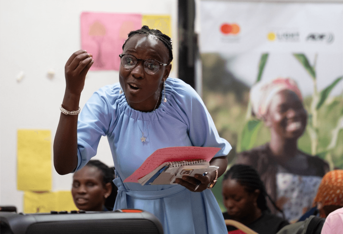 A female trainer stands and shares learnings with the programme participants. She wears glasses, a light blue dress, and holds note books in her hands. Her expression is engaging and emphatic.