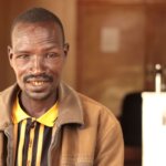 Kayoela Karata, a 35-year-old Ethiopian man, smiles and looks at the camera. He is wearing a yellow and black striped polo shirt and brown jacket.