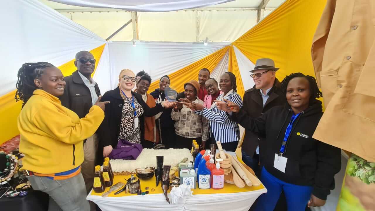 Jane Waithera, Head of the Disability Inclusion Advisory Unit at Light for the World Kenya, celebrates the award with microentrepreneurs with disabilities. There are 10 microentrepreneurs and most are pointing at the award. They are behind and to the side of a table displaying some of their work, for example with detergents, honey and crafts. 