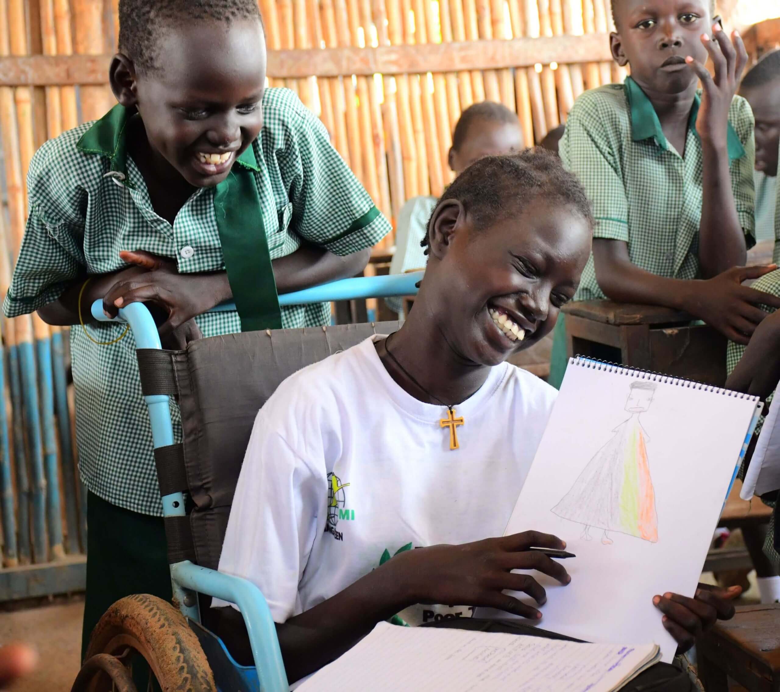 Nyaman Gatluak laughing as she shows her drawing of her friend Nyachiek to the camera. She sits in a wheelchair and is dressed in white. A young classmate leans on the back of her chair and is smiling.