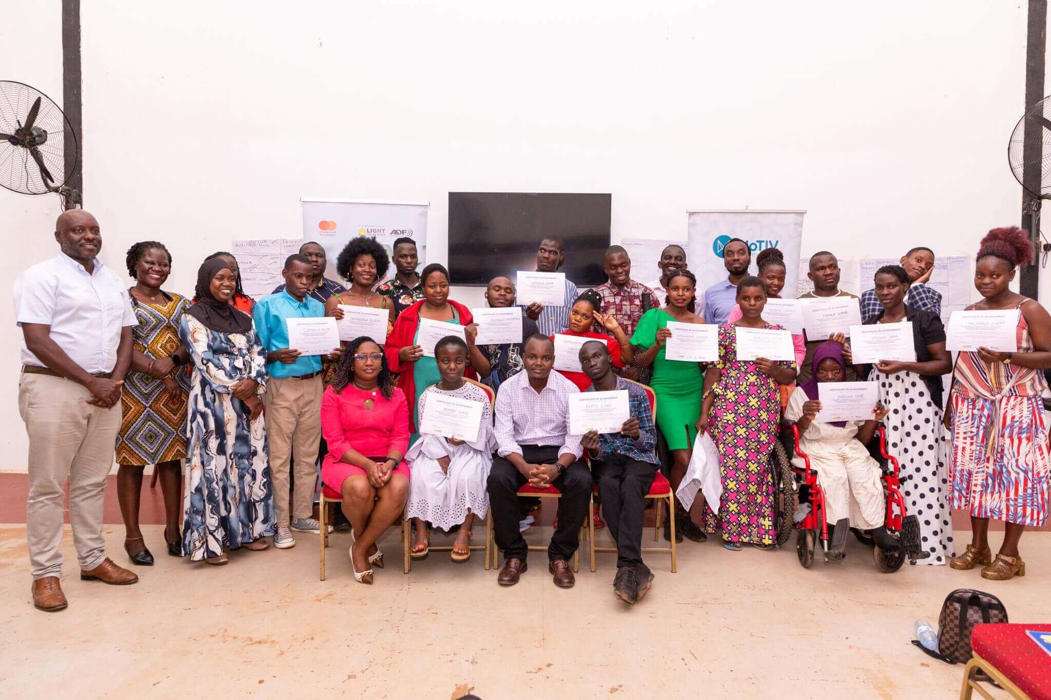 Graduates and others involved in the programme hold their graduation certificates. Some are seated and some standing in a semi-circle.