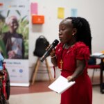 A young woman of short stature, dressed in red and holding a microphone, shares learnings from a We Can Work Academy training session.