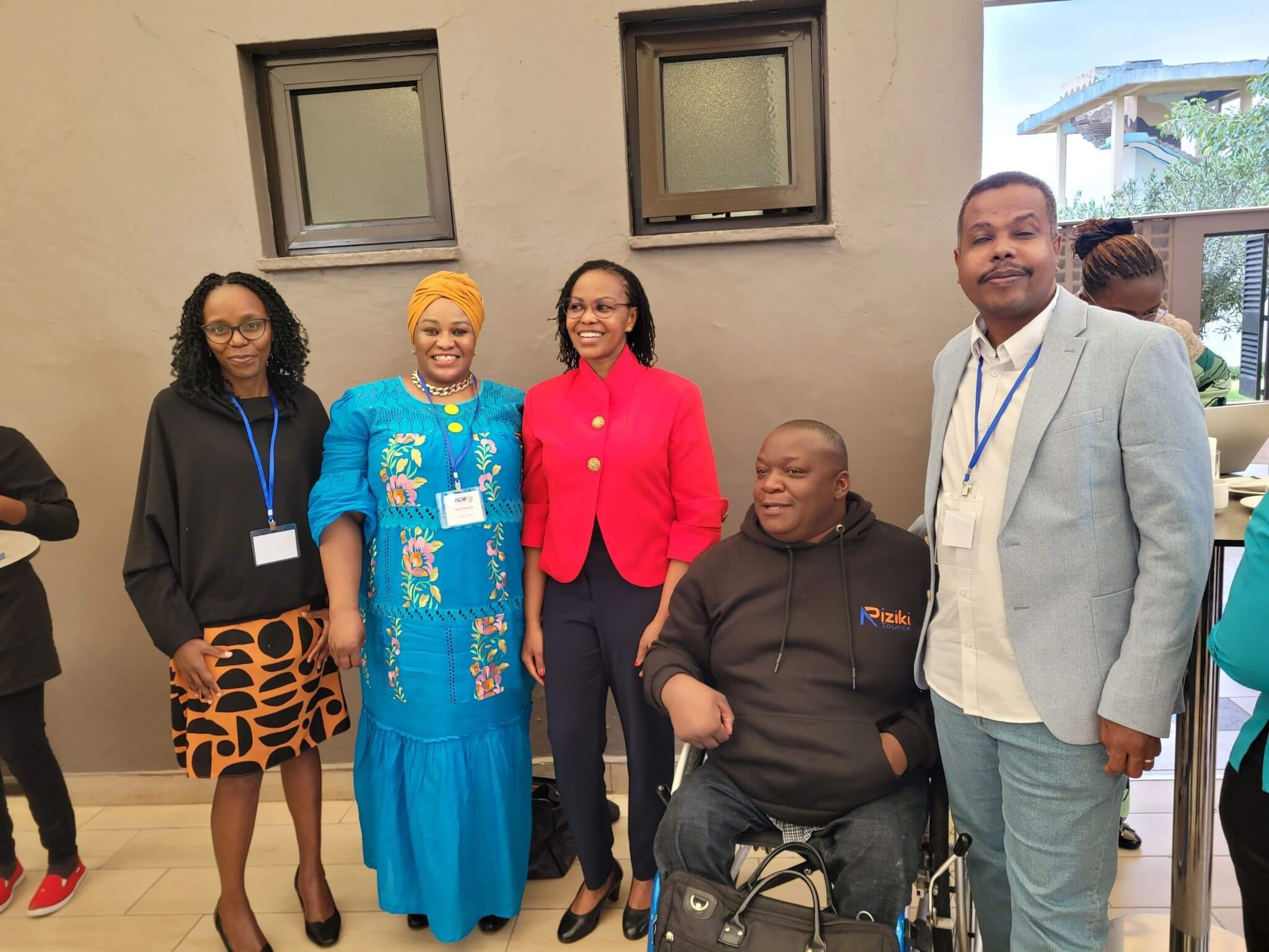 Five people are standing together. There are three womenon the left, all smiling and wearing colourful clothes. Lucy Nyaga stands in the centre wearing a formal red jacket with gold buttons. Two men are one the right. One is a wheelchair user. They standing outside in front of a grey wall with two small windows.