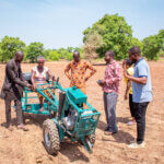 Six people are outside, in a red clay field. There are green tress in the distance. They are testing out adapted farming equipment, a woman sits on a machine with wheels.