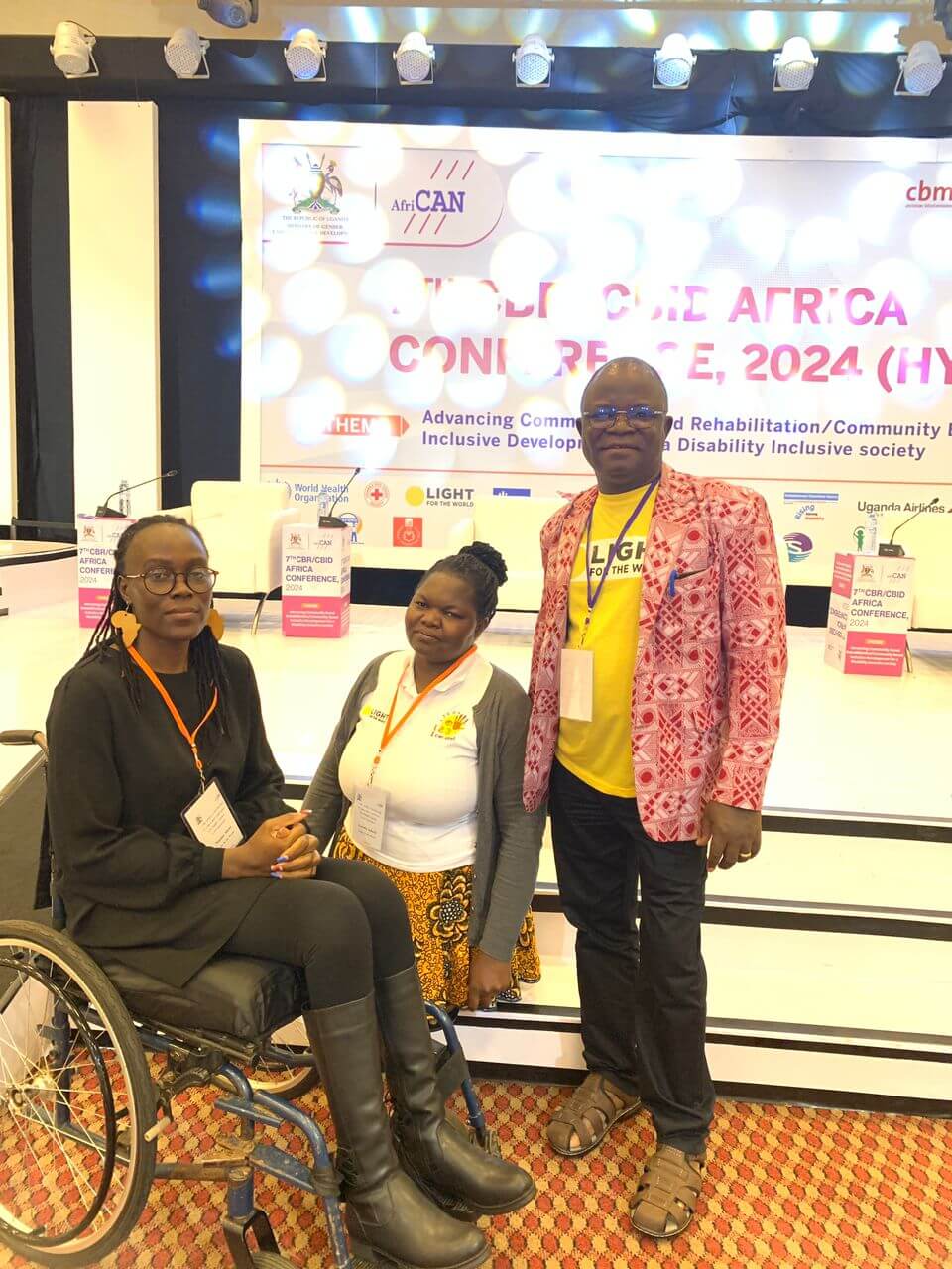 Elie Bagbila, Conny Adoch and Naome Akwee stand in front of a stage at the CBR CBID Conference. Elie is wearing a colourful red and pink jacket and Light for the World tshirt. Conny wears a colourful patterned skirt and a white poloshirt. Naome uses a wheelchair and is dressed all in black, with statement earrings in the shape of the African continent.