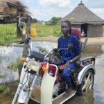 Amer Ajith, a Disability Inclusion Facilitator in Jonglei State sitting on a motorised tricycle in flood water in a village in Jonglei State.