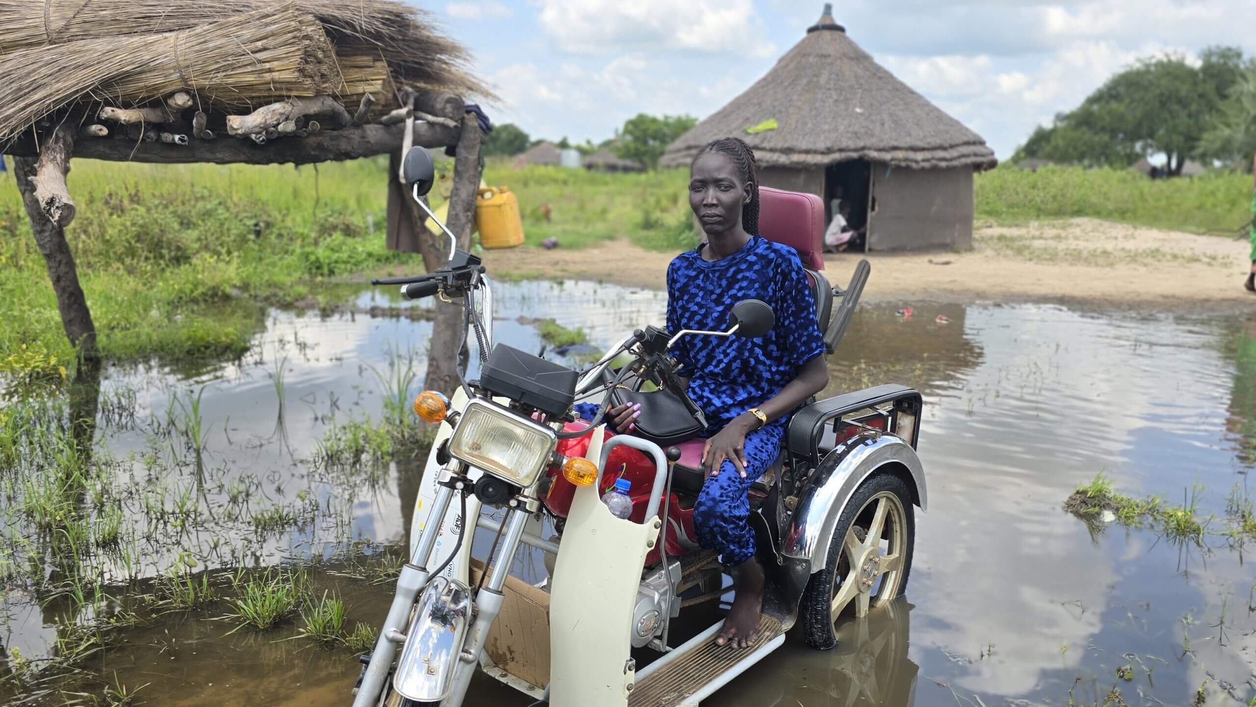 Amer Ajith, a Disability Inclusion Facilitator in Jonglei State sitting on a motorised tricycle in flood water in a village in Jonglei State.