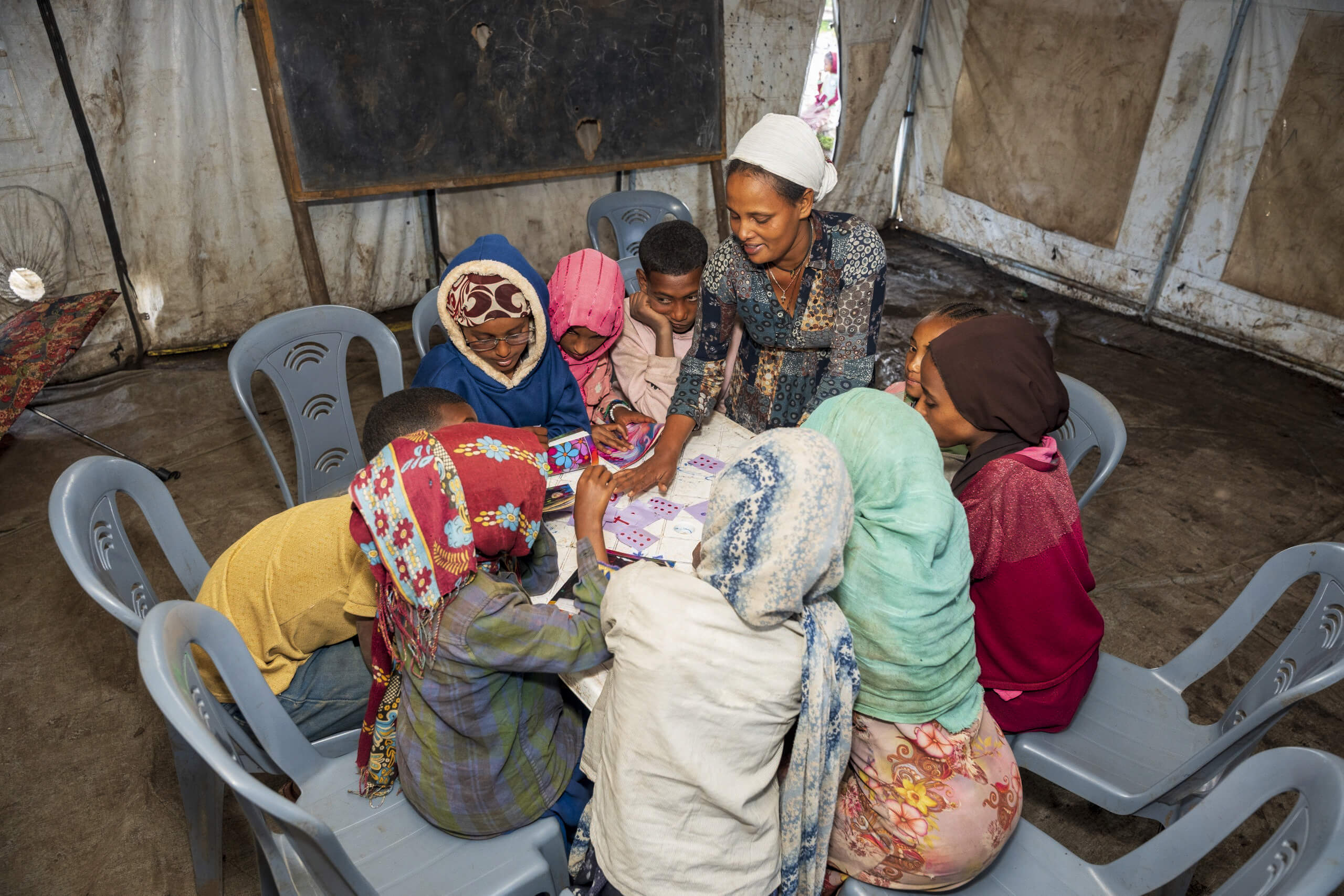 A group of children wearing colourful clothing is seated in a classroom around a table. They are playing an educational card game, receiving instructions from a woman.