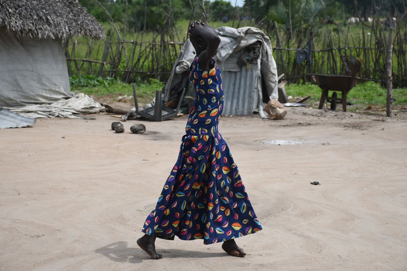 Adut, a young woman in a brightly-coloured dress, walking in her village. Her hands are on her head and her face is not visible. 