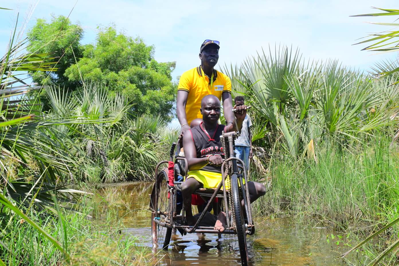 Ayuen, a farmer and church leader, in a tricycle in flood water. He is pushed by Daniel Anyang Disability Inclusion Facilitator with Light for the World.