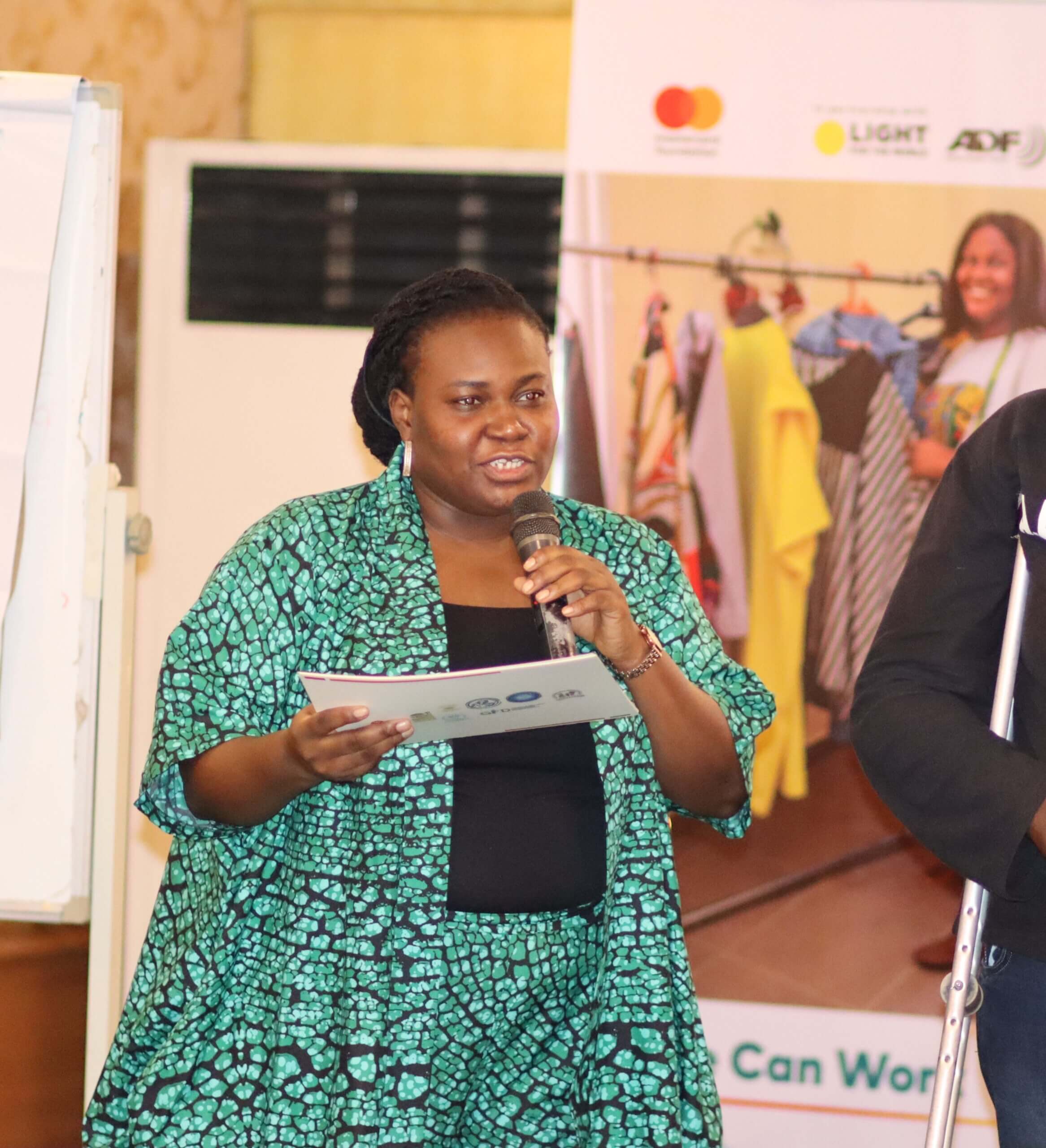  Adedotun Esan stands with a microphone, speaking to a group of people. She is wearing a turquiose and black patterned suit and black top. She looks engaging and is wearing big silver earrings and holding some papers.