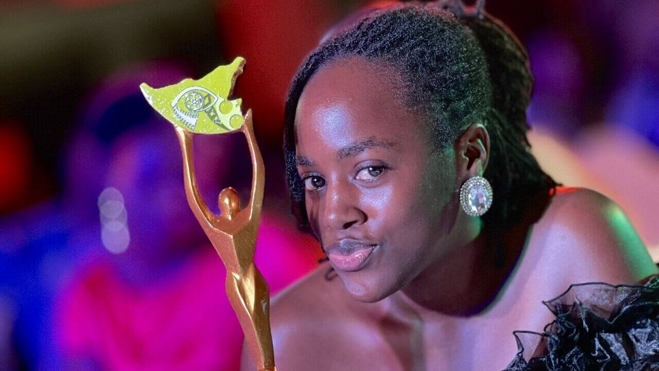 A woman wearing earings and a party dress is holding an a golden award tropy. © Doreck Ankunda / Light for the World