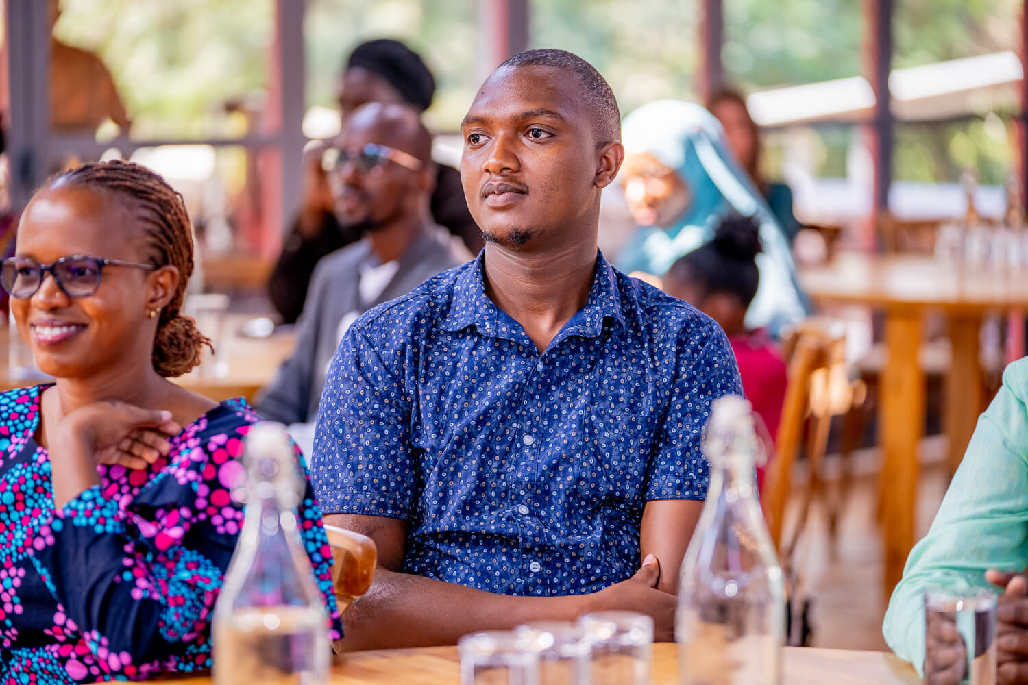 A man wearing a blue patterned shirt, Wilson Kamau, sits in a room with other participants. He has corssed his armes and is following the action outside of the image attentively.