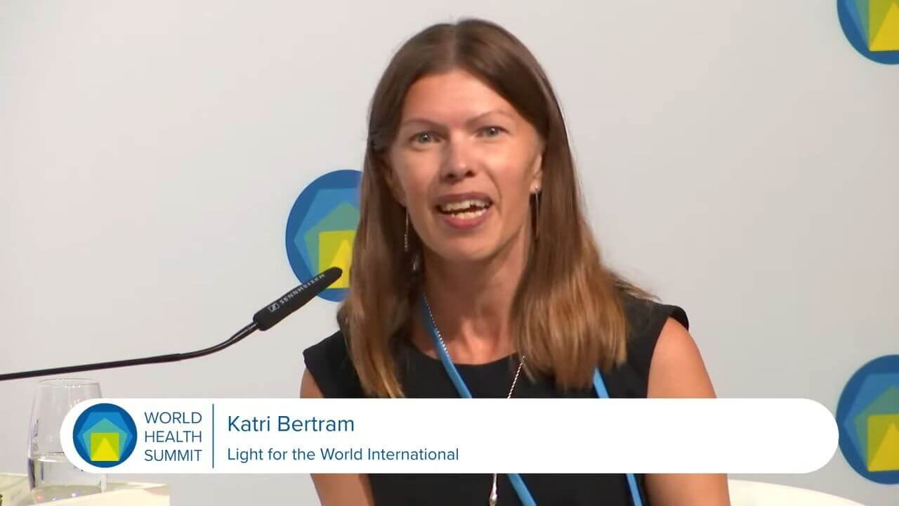 A woman with long brown hair, Katri Bertram, is moderating a panel at the World Health Summit in Berlin. She speaks towards the camera and uses a microphone.