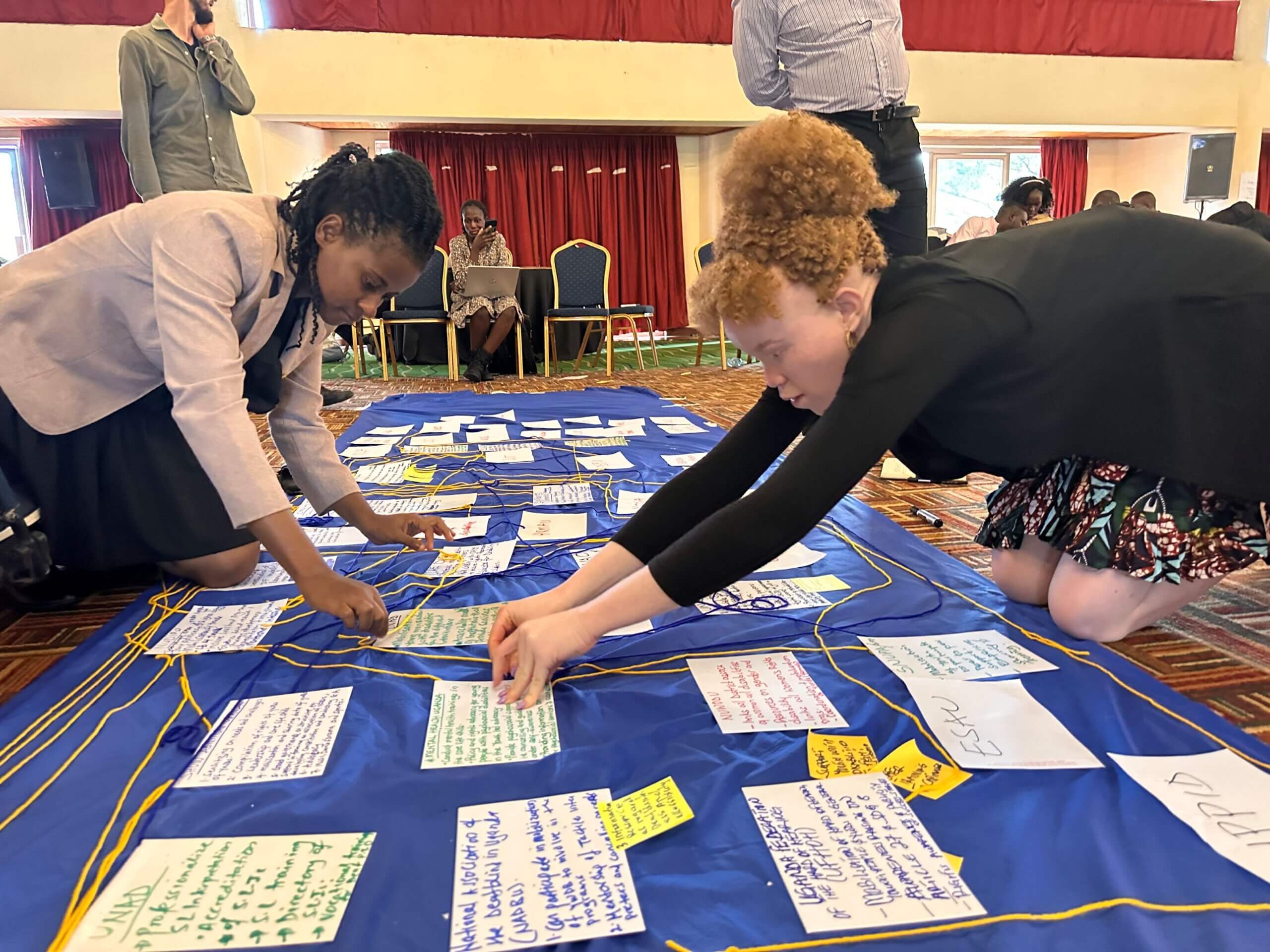 Young women take part in a deep dive brainstorm session during co-creation week in Uganda. They are adding cards with writing to a blue canvas.