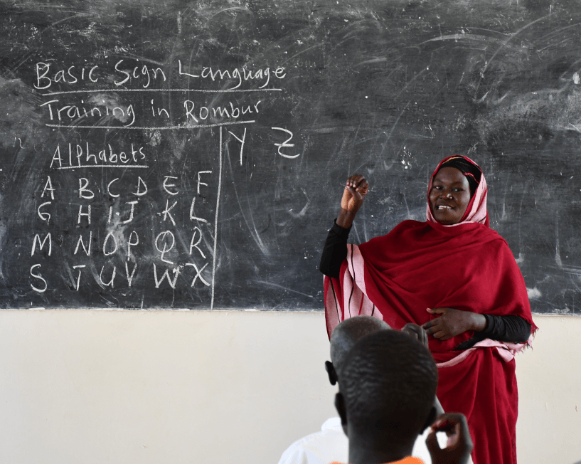 Hanadi, a trainee teacher, practises signs during a class. She is in front of a blackboard and wearing a red dress.