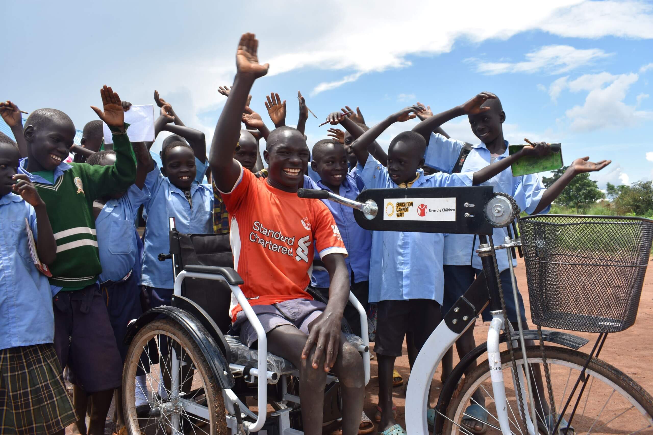 Moses on his new tricycle surrounded by his classmates. Moses has his arm raised and is smiling. His classmates are also smiling.