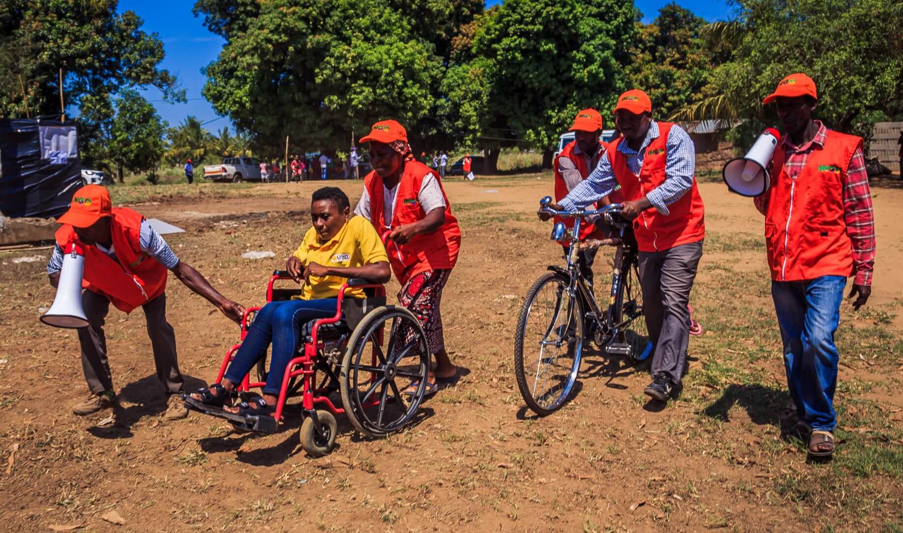 People take part in a Cyclone simulation exercise in Buzi district. Helena Rego, a DIF who uses a wheelchair, is supported by two people in red high-visibility jackets and hats. Three other men are also in high-vis clothing. One has a megaphone.