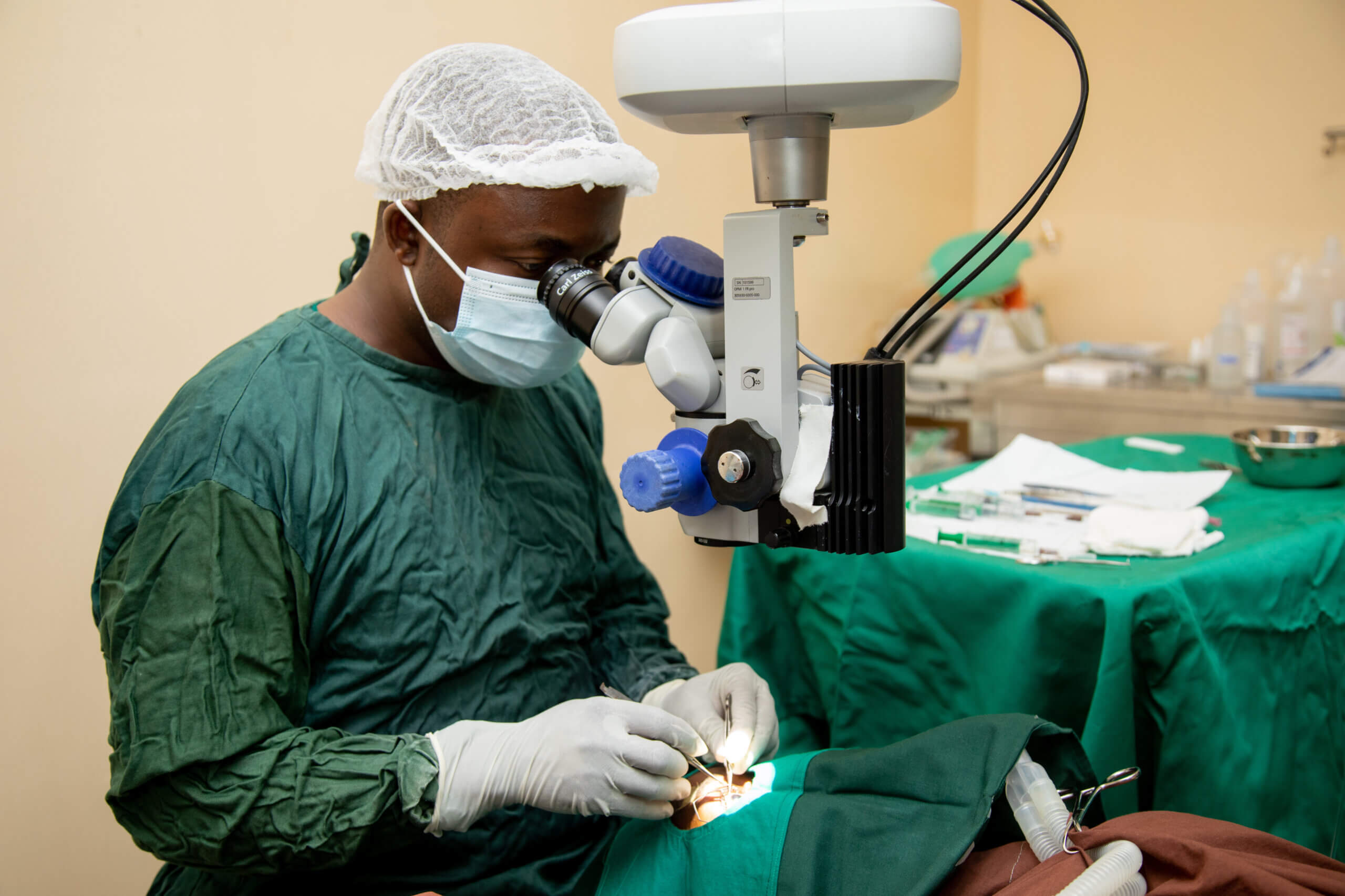 Dr. Vasco da Gama performs a surgery. He is wearing white medical gloves, a cap and face mask and a green medical slip and looks into surgical equipment. The patient lies on a table, with everything covered but one eye, where medical tools can be seen.