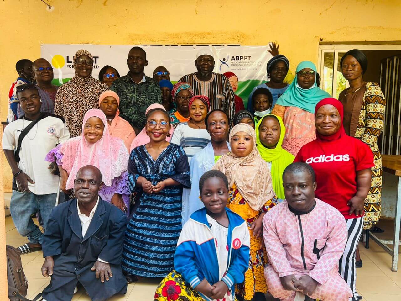 A group of about 30 people sit and stand in a group photo. The group is made up of Light for the World, Burkinabè Association of Short Stature Persons and persons of short stature. Everyone wears colourful clothes and is smiling.