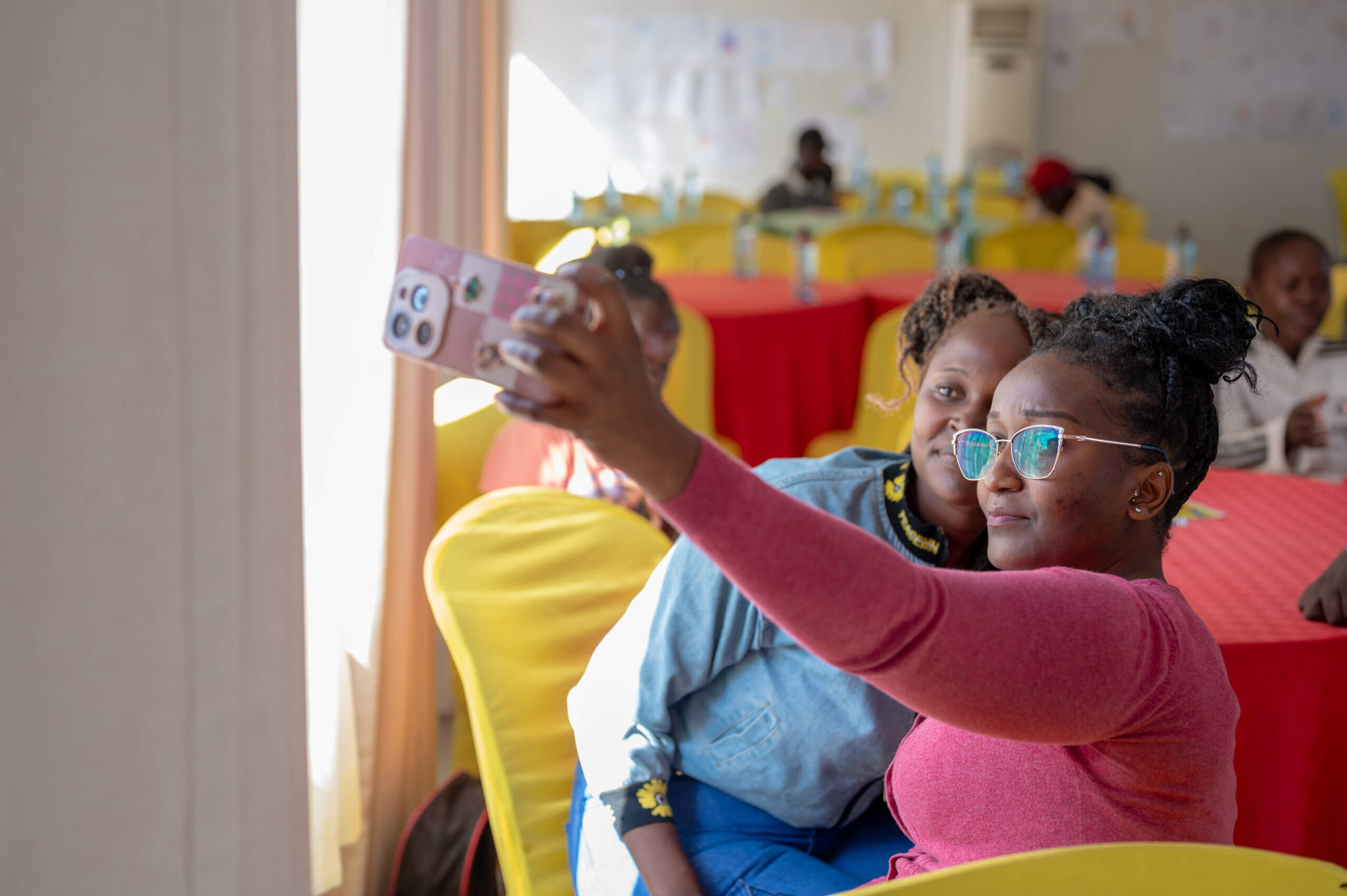 Two girls pose for a selfie. One is wearing glasses and a pink tshirt, while the other wears a blue shirt and trousers. They both look serious but happy.