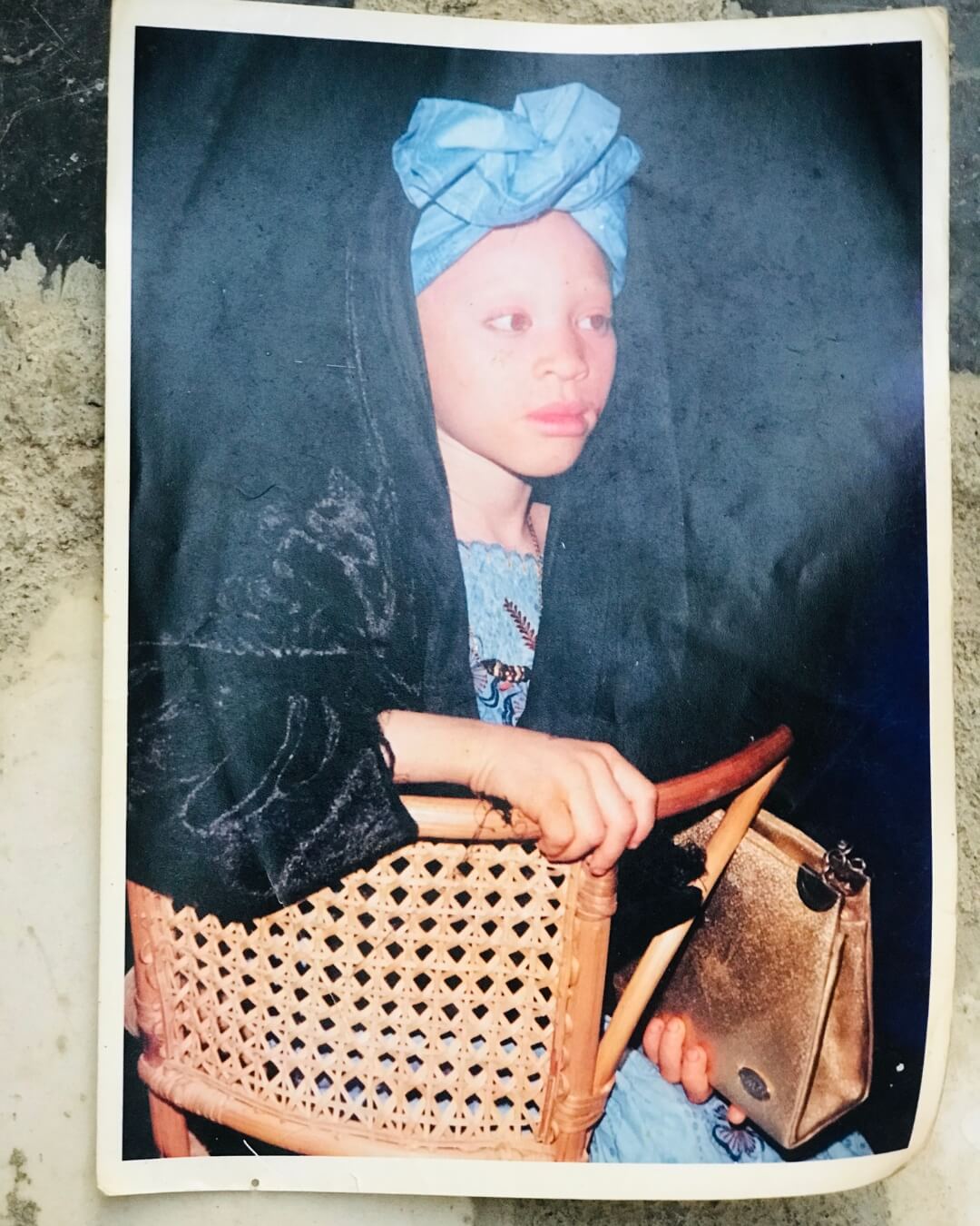 The image is of a printed picture. A young girl smiles softly, sitting on a wooden chair. She is wearing a bright blue head wrap and blue dress, covered by a black layer. The image is slightly dated and looks as if it captures a lot of memories.