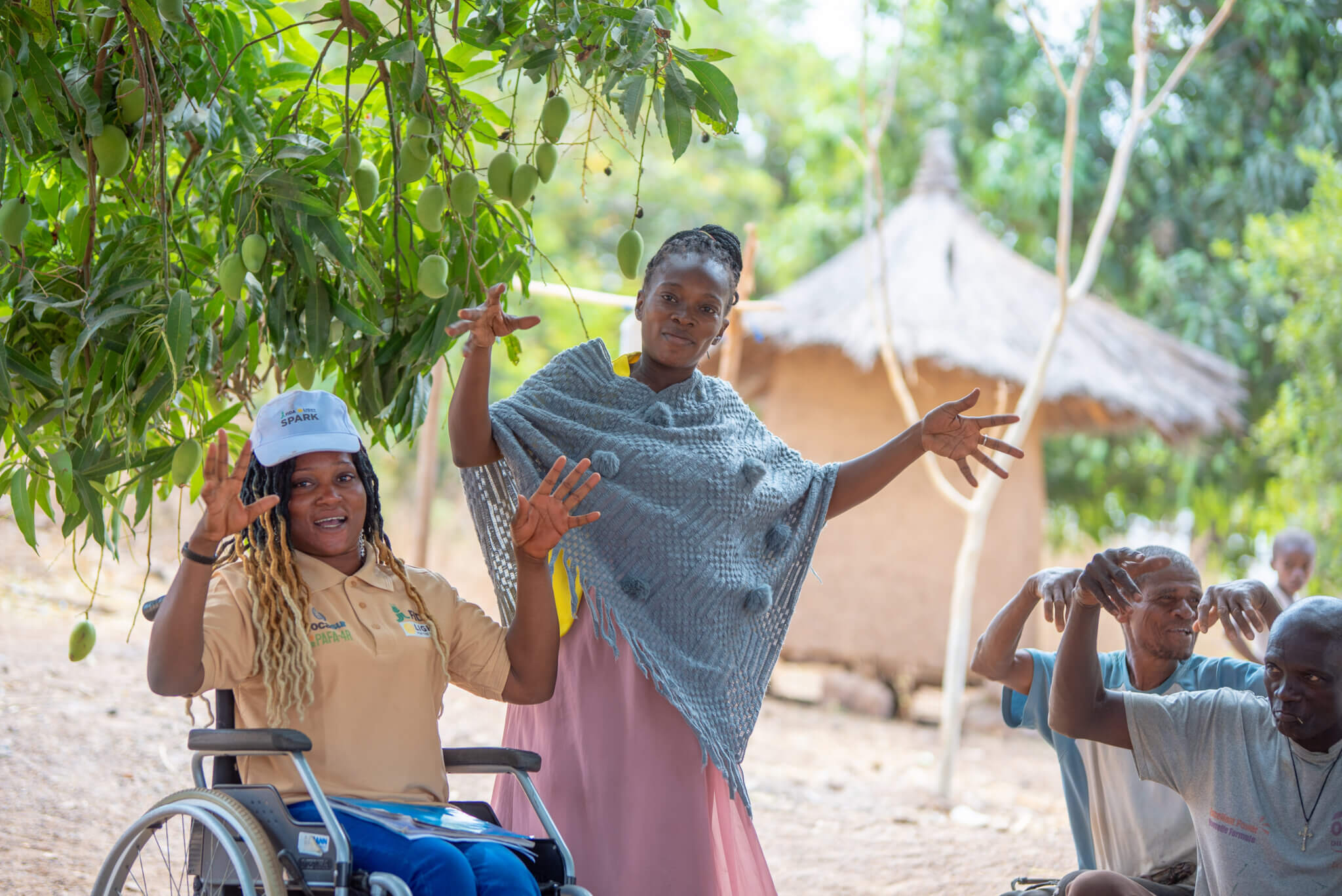 Two women are outside with hands raised to the camera. It is lush and green and there is a tree next to them. One woman wears a Light for the World top and cap and sits in a wheelchair. The other woman wears a blue poncho and pink skirt. They both look playful and are softly smiling. There are men to the right in the background also with their hands raised.