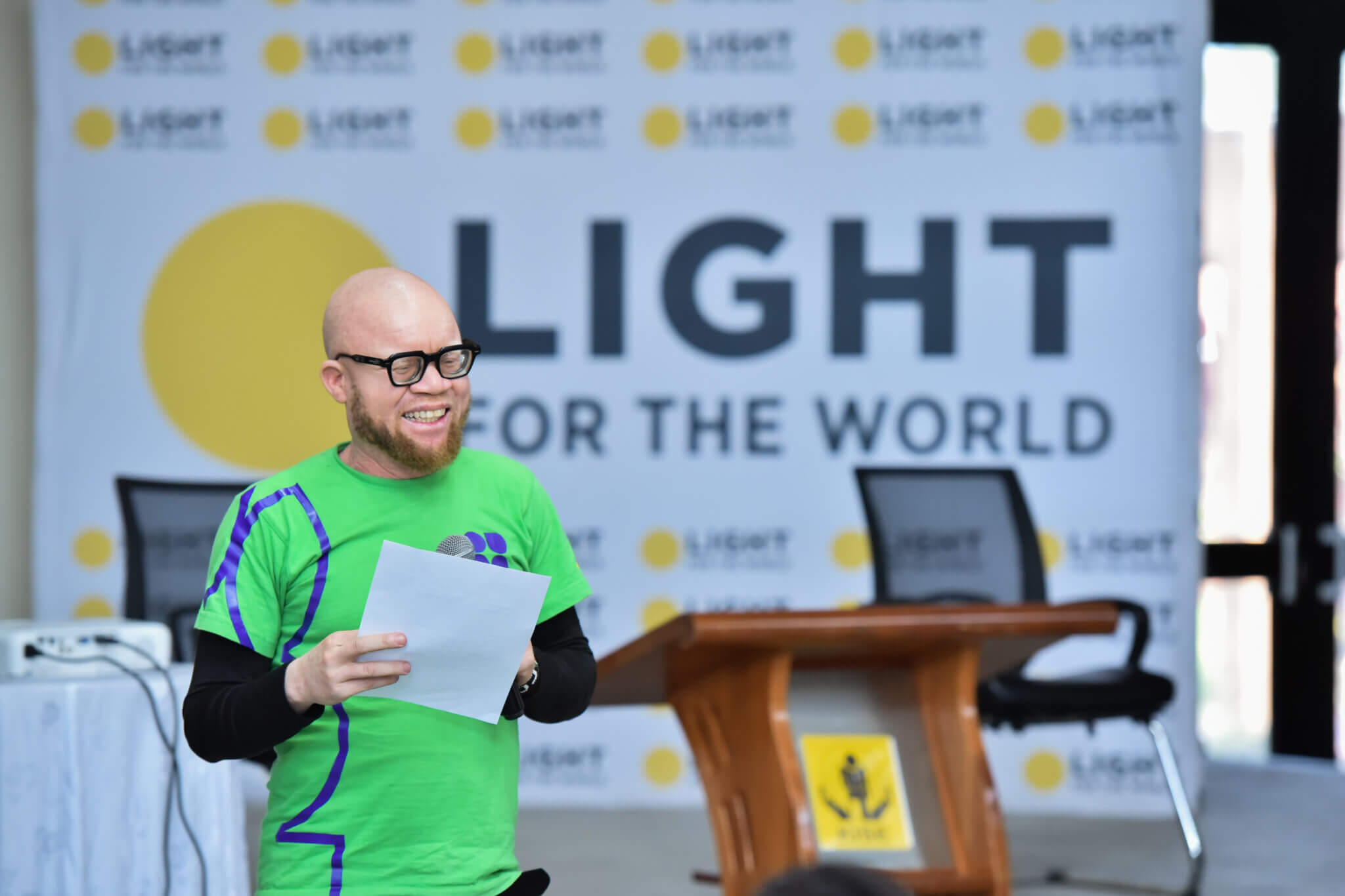 A man in a green shirt speaks on a stage. He is wearing black glasses and holding a piece of paper. The Light for the World logo appears behind him.