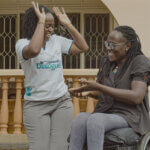 Two women are outside smiling and high fiving with all hands. Doreck stands up wearing a white tshirt and Naome sits in a wheelchair with a grey shirt and leggings. You can tell they are good friends and having fun together,