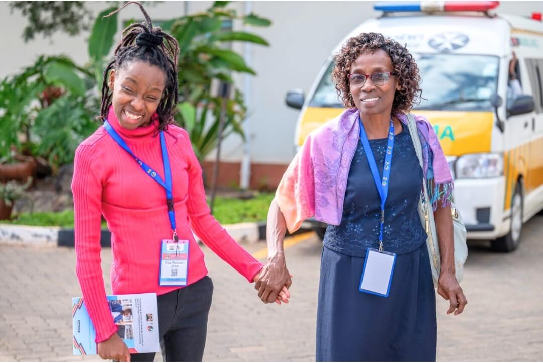 Two women stand together holding hands. They are both wearing pink and purple colourful clothes and blue lanyards around their necks. The woman on the left, Elsa, has a big grin, whilst the woman on the right smiles and looks into the camera. They are standing outside, in front of some plants and a large van.