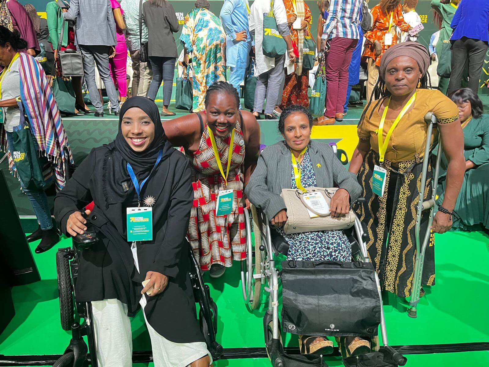 Four women smile in a photo. They are in a green conference room. Two women sit in wheelchairs and the other two stand alongside. They are all wearing colourful clothes and smiles on their faces.