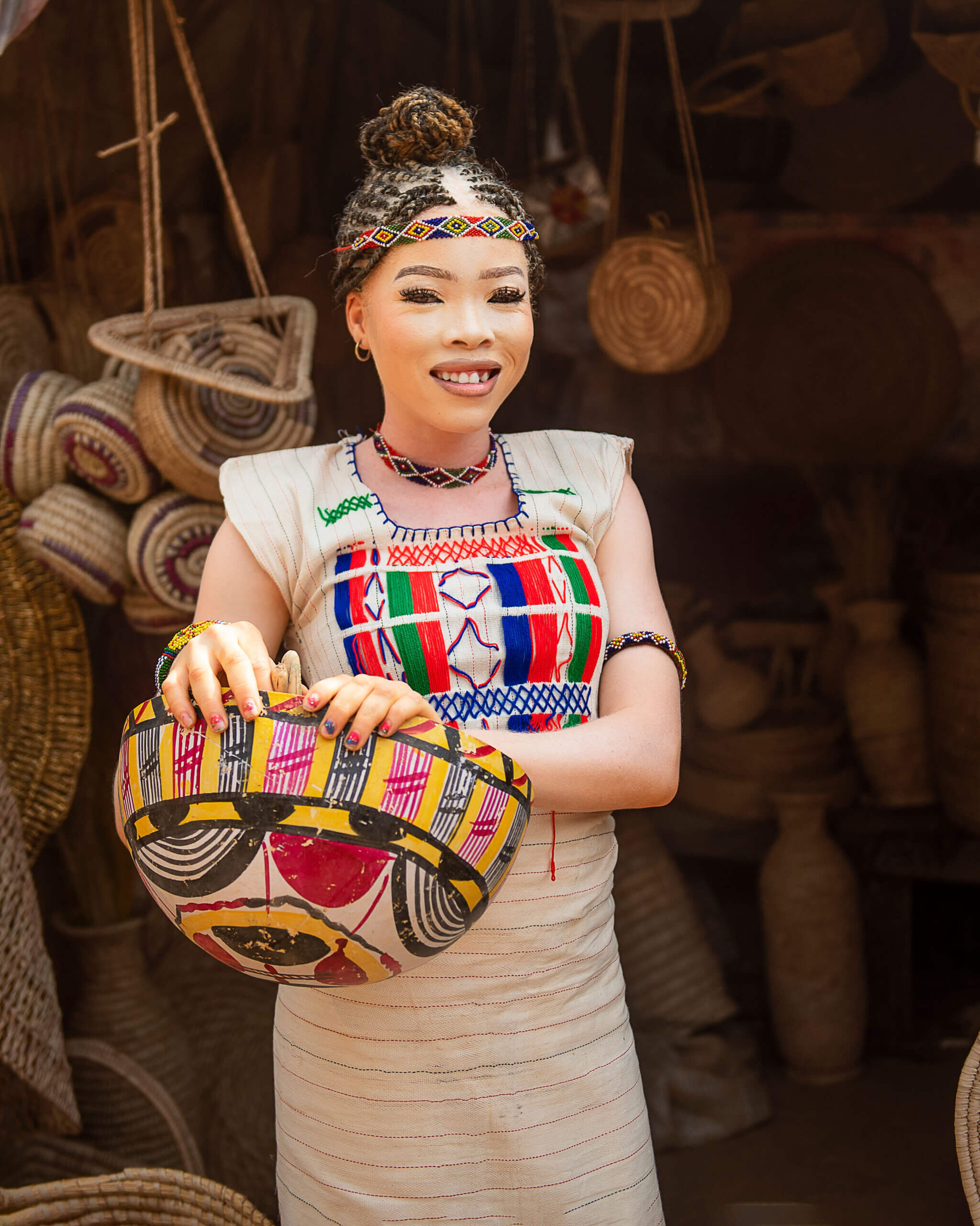 A woman stands at a market place in from of lots of brown woven baskets. She is smiling and wearing a beautiful colourful patterned dress and headband. She has her hair in brainds up, and has colourful painted nails and is holding a colourful basket.